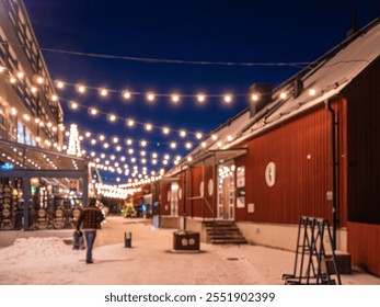 Deliberately out of focus, a cozy winter evening scene with string lights illuminating a snowy street between red wooden buildings. A person walks along the path, creating a festive atmosphere. - Powered by Shutterstock