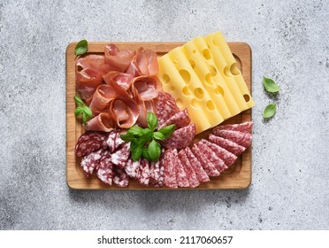 Delicacies. Cheese, prosciutto, salami on a wooden square board on a concrete background. - Shutterstock ID 2117060657