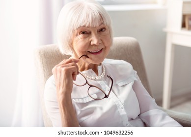 Delicate beauty. The close up of a pretty elderly woman posing for the camera, having removed her eyeglasses, while sitting in an armchair