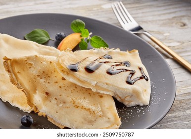 Delicate crêpes filled with cream, drizzled with chocolate sauce and garnished with fresh mint, a peach slice, and blueberries. Served on a black plate with powdered sugar. - Powered by Shutterstock