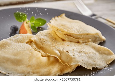 Delicate crêpes filled with cream, drizzled with chocolate sauce and garnished with fresh mint, a peach slice, and blueberries. Served on a black plate with powdered sugar. - Powered by Shutterstock