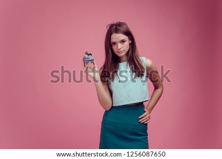 delicate pink background in the new fashion photo art studio and a model with long brown hair, big eyes, nice figure holds a cream cup cake with figs in here hand.  dressed in green top and skirt.