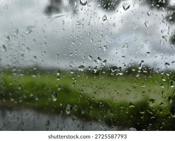 Delicate raindrops rest on a window pane, creating a soft, blurred veil over the lush green fields and overcast sky