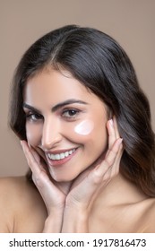 Delicate texture. Close up face of smiling beautiful woman with cream on cheeks and hands near cheeks
