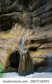 A delicate waterfall threads through weathered stone carved by centuries of water flow, cascading over mineral-stained rock walls adorned with vibrant green moss.