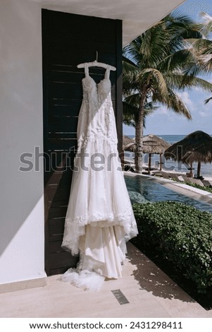 A delicate wedding dress hangs on a hanger outdoors against the background of the ocean and palm trees. Tropical wedding in a hotel near the ocean, the sea. Long white wedding dress.