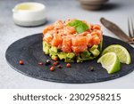 Delicious avocado and raw salmon salad, tartare, served on a black plate with lime, light background