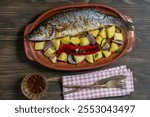 Delicious baked big sea bass fish with spices in a clay plate and fried vegetables, close up, top view. Cooked sea bass with potatoes, red chili pepper, onion and garlic