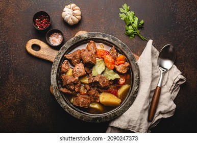 Delicious beef meat stew dish with potatoes, carrot and gravy in rustic vintage metal bowl with spoon, bunch of fresh parsley, garlic cloves, spices on brown concrete background top view flat lay - Shutterstock ID 2090247373