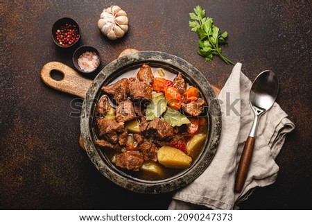 Delicious beef meat stew dish with potatoes, carrot and gravy in rustic vintage metal bowl with spoon, bunch of fresh parsley, garlic cloves, spices on brown concrete background top view flat lay