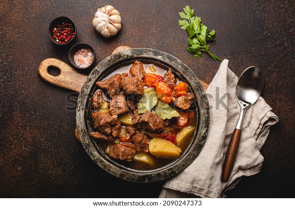 Delicious beef meat stew dish with potatoes, carrot\
and gravy in rustic vintage metal bowl with spoon, bunch of fresh\
parsley, garlic cloves, spices on brown concrete background top\
view flat lay