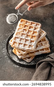 Delicious belgian waffles in stack on dark background. Woman hand is sprinkling waffles with powdered sugar close up. Vertical