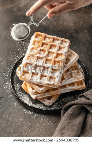 Delicious belgian waffles in stack on dark background. Woman hand is sprinkling waffles with powdered sugar close up. Vertical