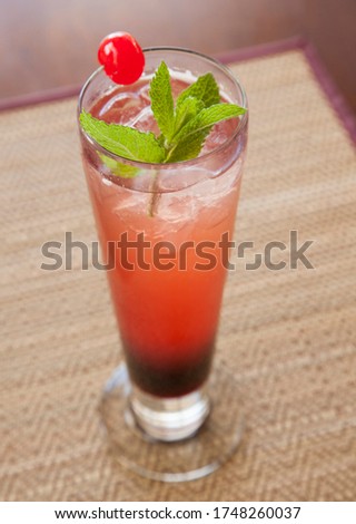 A delicious berry cocktail in a high glass with ice, cherry and mint garnish on a placemat in a restaurant.
