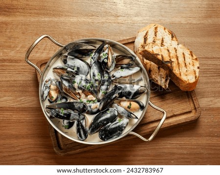Delicious black mussels in creamy cheese and garlic sauce. Mediterranean hot black mussels in sauce cooked in metal pan on wooden cutting board with grilled bread on wooden background