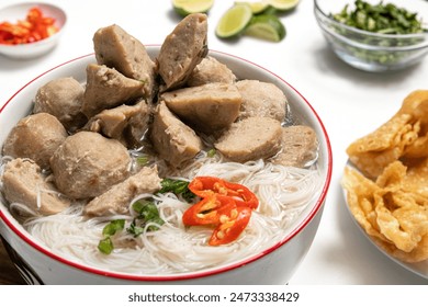 Delicious bowl of Bakso Solo meatballs, rice noodles, and fresh herbs, surrounded by various garnishes lime, chili, greens, and crispy wontons, on a wooden tray with a white background - Powered by Shutterstock