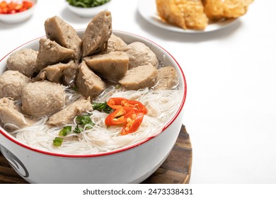 Delicious bowl of Bakso Solo meatballs, rice noodles, and fresh herbs, surrounded by various garnishes lime, chili, greens, and crispy wontons, on a wooden tray with a white background - Powered by Shutterstock