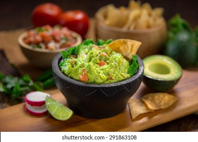 A Delicious Bowl Of Guacamole Next To Fresh Ingredients On A Table With Tortilla Chips And Salsa
