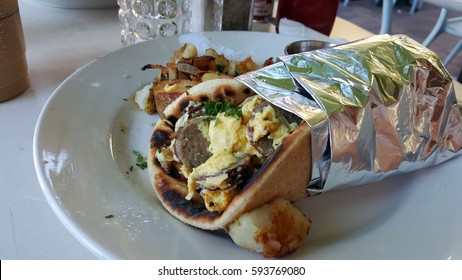 A Delicious Breakfast Burrito At An Outdoor Cafe, Wrapped In Foil With Eggs, Sausage, And Cilantro In A Pita Wrap, With Breakfast Potatoes And Coffee In The Background