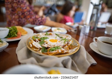 Delicious Chilaquiles, Cup Of Coffee, And More Dishes On Brown Table With Blurry Family Taking A Picture As Background. Woman And Little Girl At Restaurant Table Ready To Eat. Authentic Mexican Food