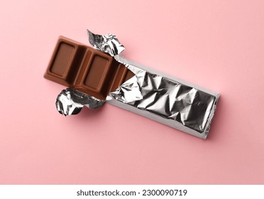 Delicious chocolate bar wrapped in foil on pink background, top view