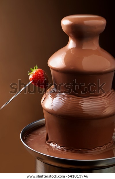 Delicious chocolate fountain fondue with a ripe\
red strawberry on a fork dripping the sauce after being dipped and\
copy space behind