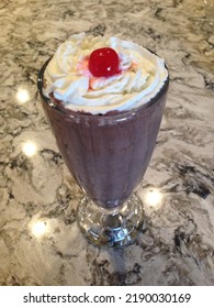 Delicious Chocolate Milkshake With Whipped Cream And A Cherry On Top In A Milkshake Glass With A Granite Countertop Background. 