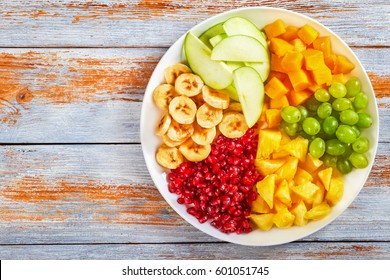 Delicious Colorful Fruit Salad Set - Banana, Apple, Mango, Pineapple Cut In Slices, Grapes And Seeds Of Garnet On White Plate, On Old Peeling Paint Boards, View From Above