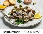 Delicious cooked sea escargo snails with herbs, butter, garlic on metal plate with forks. wine glass. gourmet food. Restaurant menu, Traditional French cuisine,