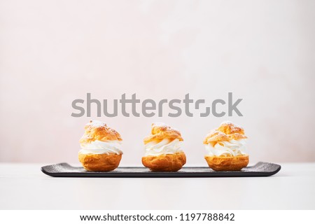 Delicious cream puff cakes with cream and powdered sugar on a black plate on white marble table over pink background. Selective focus, copy space.