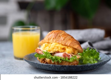 Delicious croissant sandwich with ham, cheese and scrambled eggs on plate served with glass of orange juice