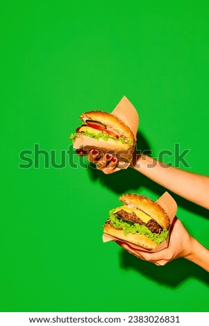 Delicious fast-food. Hands holds appetizing hot hamburger and cheeseburger in paper against vivid green background. Concept of junk food, menu, delivery, catering, take away. Copy space. Ad