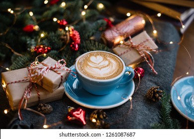 Delicious fresh festive morning cappuccino coffee in a ceramic blue cup on the warm cover with little wrapped gifts, red ornamentals, fireflies and spruce branches - Powered by Shutterstock