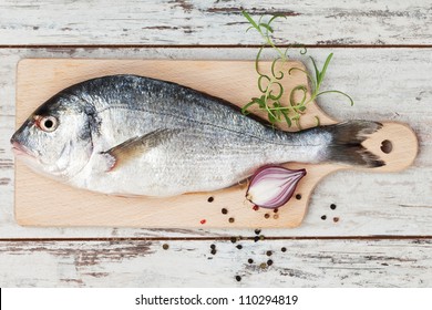 Delicious fresh sea bream fish on wooden kitchen board with onion, rosemary and colorful peppercorns on white textured wooden background. Culinary healthy cooking. - Shutterstock ID 110294819