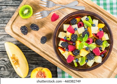 Delicious Fruit And Berry Summer Salad Decorated With Mint Leaves On Clay Dish On Cutting Board With Dessert Forks, Half Of Peach And Slice Of Melon On Rustic Boards, View From Above