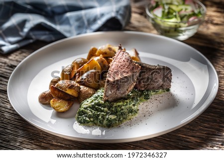 Delicious grilled beef sous vide steak served on a white plate with crunchy roasted potatoes, spinach dip and fresh leaf lettuce salad. 