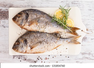 Delicious grilled sea bream fish on kitchen board with rosemary, lemon and colorful peppercorns on white textured wooden background. Culinary healthy cooking.