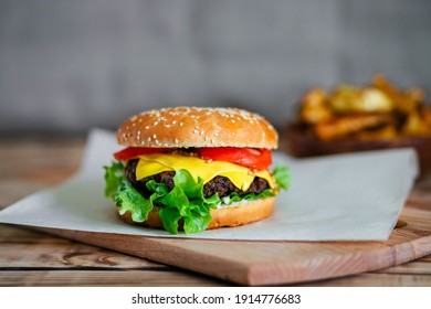 A delicious homemade cheeseburger with French mustard sits on the chopping board. On the background, the potato slices are blurred