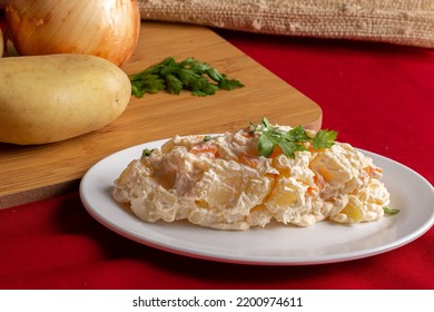 A delicious homemade creamy potato salad served on a white plate on a table with a red tablecloth and fresh ingredients in the background. 