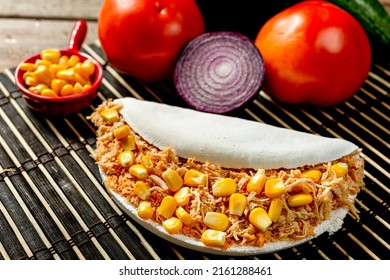 Delicious homemade tapioca stuffed with cooked and shredded chicken with boiled green corn, selective focus, with purple onion and red tomatoes in the background on a rustic wooden table