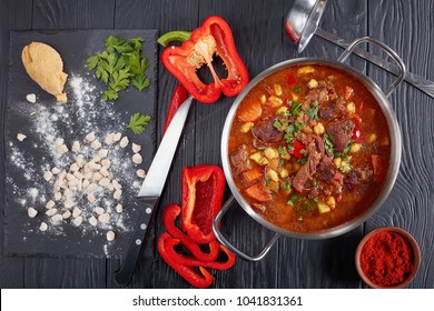 Delicious Hot Hungarian Goulash With Beef Meat, Paprika, Vegetables, In A Pot. Raw Csipetke With Dough On Board, Sweet Pepper And Soup Ladle On Table, Classic Recipe, View From Above