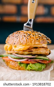 Delicious Juicy Burger Close Up From Brioche Bun. Chicken Steak, Cheese, Onion, Tomato, Cucumber, Lettuce, Stuck Knife, Sauce. Cafe Table. Wooden Board