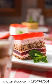 A Delicious Looking Dessert Cake Served On A White Plate. Layered Sponge Cake With Jello Layers And Mint Leaf On Top. Fruit Berry Dessert. On A Rustic Wooden Table With Red Plaid Tablecloth.