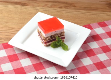 A Delicious Looking Dessert Cake Served On A White Plate. Layered Sponge Cake With Jello Layers And Mint Leaf On Top. Fruit Berry Dessert. On A Rustic Wooden Table With Red Plaid Tablecloth.