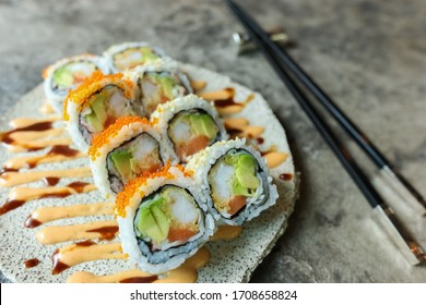 Delicious Maki With Raw Salmon, Crab Meat, Avocado Topped With Orange Tobikoon On The Ceramic Plate. Japanese Sushi Rolls On Stone Table. Front Two Are In Focus, The Rest Are Blurry. Image