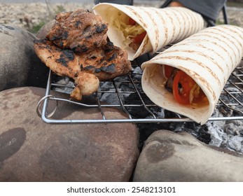 A delicious meal is being cooked outdoors with grilled chicken resting on a wire rack over hot coals. Fresh wraps filled with vegetables are being prepared by the river on a sunny day. - Powered by Shutterstock