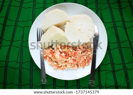 A delicious meal of boiled yam and fried scrambled eggs prepared with sliced onions, tomatoes and red pepper. Served in a white plate on a colorful green pattern table cloth with a knife and fork 