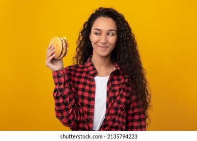 Delicious Meal. Portrait Of Hungry Female Model Holding Looking At Burger In Hand. Smiling Casual Woman Enjoying Tasty Fast Food, Eating Snack Isolated Over Orange Yellow Studio Wall, Banner