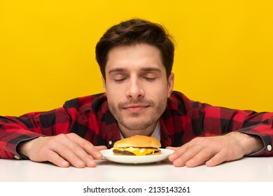 Delicious Meal. Portrait Of Hungry Male Model Eating Burger. Smiling Millennial Man Smelling Tasty Fast Food On A Plate With Closed Eyes Sitting At Table, Isolated Over Orange Yellow Studio Background