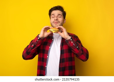 Delicious Meal. Portrait Of Hungry Male Model Eating Burger. Smiling Casual Man Holding And Smelling Tasty Fast Food, Chewing Snack With Closed Eyes Isolated Over Yellow Studio Background Wall